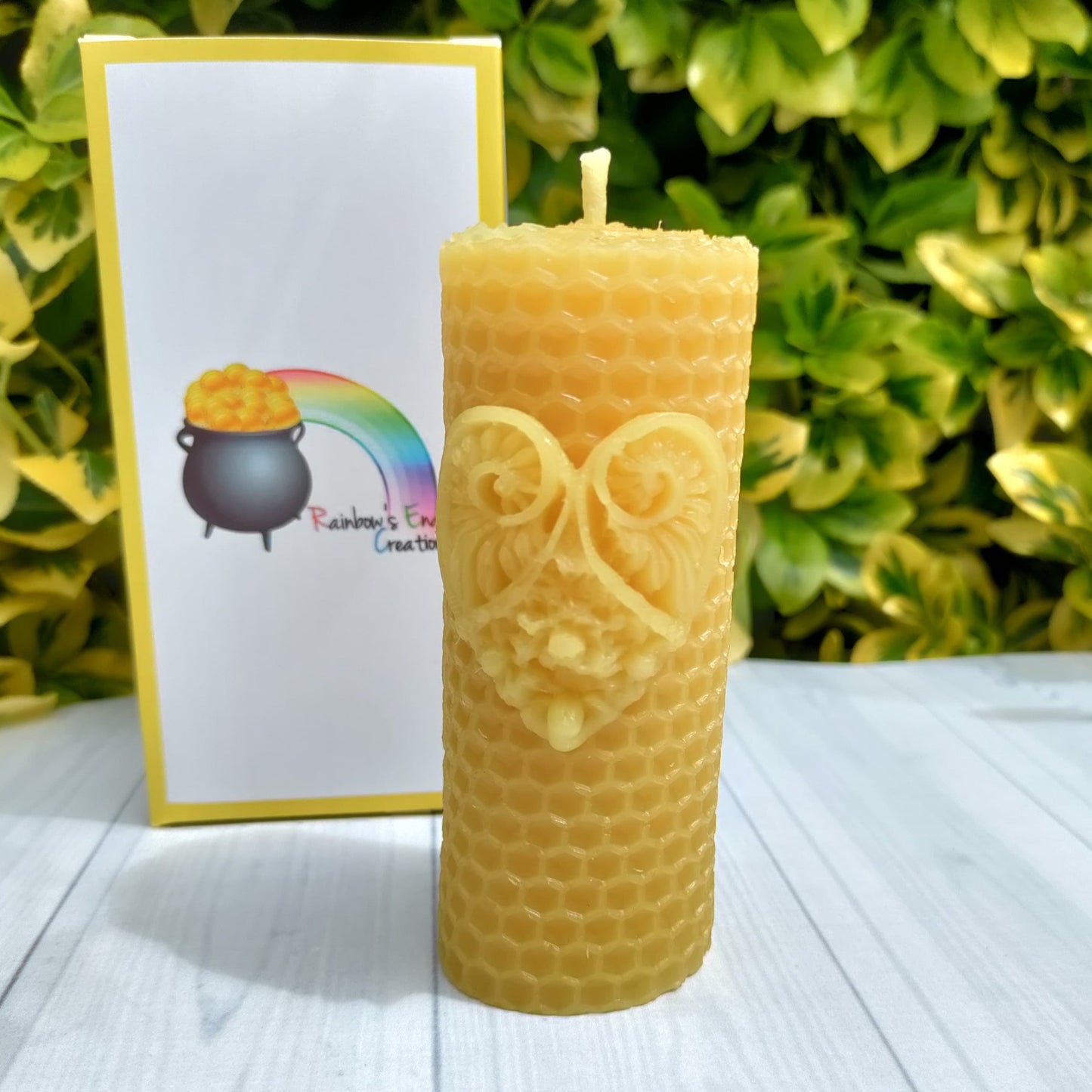 a pillar candle that looks like a beeswax sheet with the hexagon pattern on it but is a solid candle. Has a heart motif on the front. Standing in front of a white gift box with yellow edging and the rainbows end creations logo on the front.