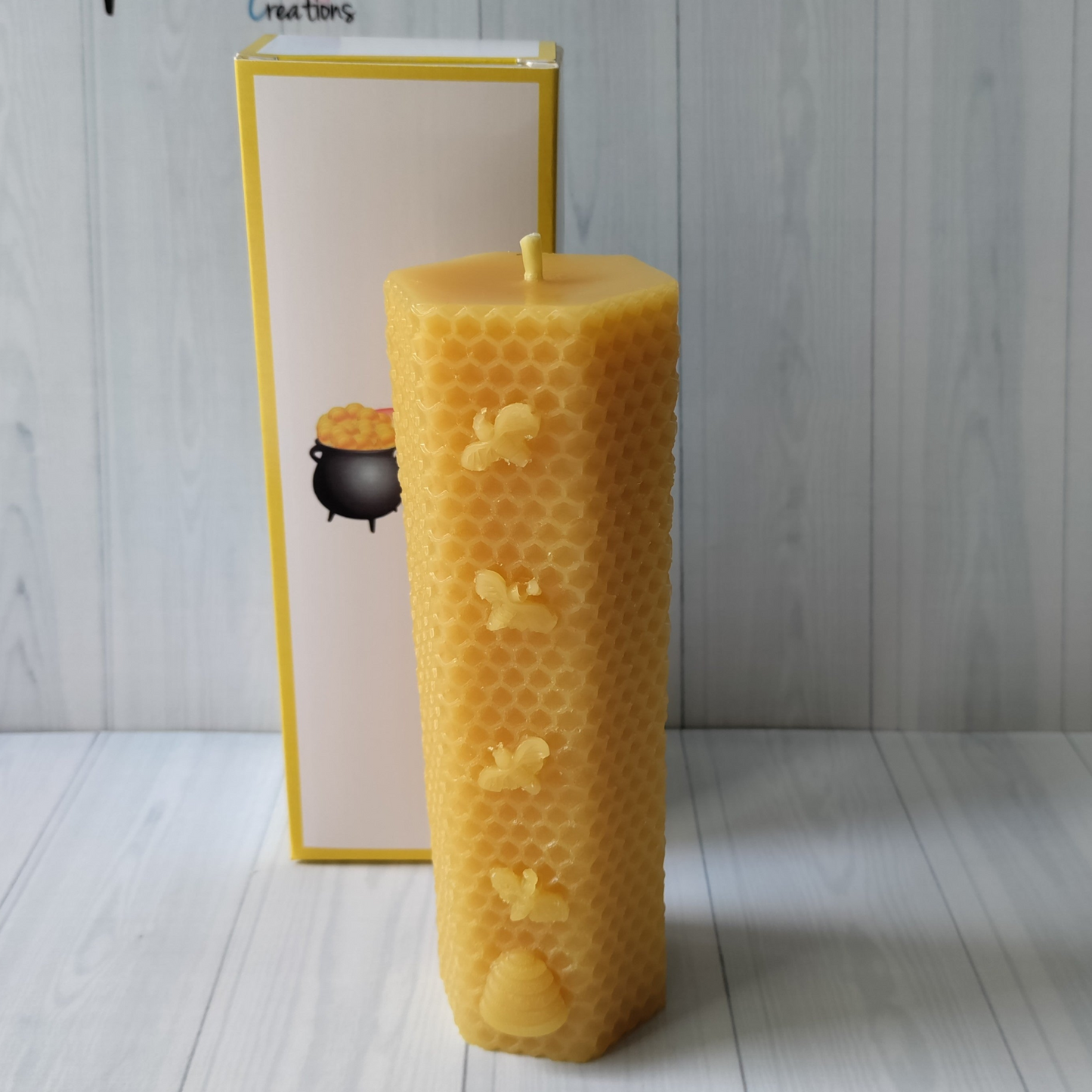 a tall hexagonal shaped golden coloured candle, It has a honeycomb pattern on the outside with four small bees and a beehive on the front. Standing in front of a white gftbox with yellow edging and the rainbows end creations logo on the front.