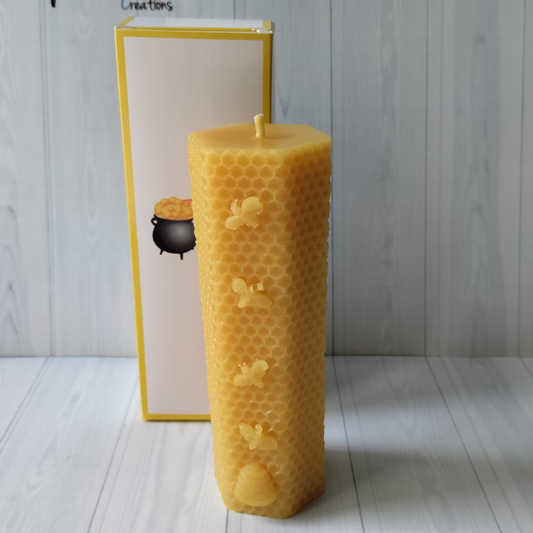 a tall hexagonal shaped golden coloured candle, It has a honeycomb pattern on the outside with four small bees and a beehive on the front. Standing in front of a white gftbox with yellow edging and the rainbows end creations logo on the front.